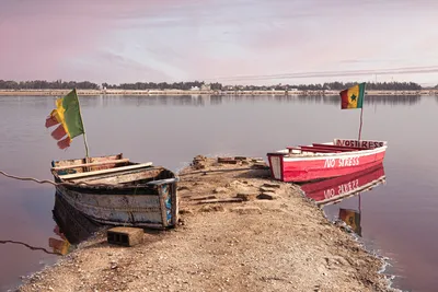 Two boats with Senegalese flags moored at a natural jetty on the Pink Lake, Senegal, under a pastel sunset sky