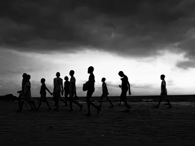 High-contrast black and white silhouette of a group of children walking along the bright horizon line between a dark sky and sea at Toubab Diallaw beach, Senegal