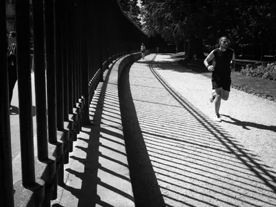 High-contrast black and white photo of a jogger on a gravel path in the Luxembourg Gardens, Paris, with long shadows cast by the park's large gates