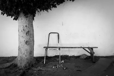 Black and white still life of an improvised bench made from a wooden plank resting on a broken chair in the shade of a tree, Dakar