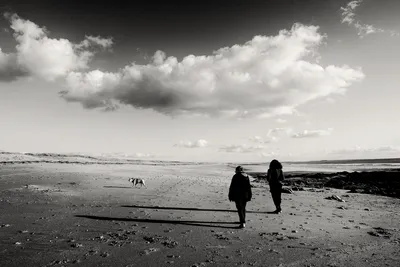 Wide black and white shot of two women walking with a dog on Plouhinec beach in Brittany under a large white cumulus cloud