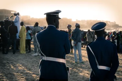Two police officers in uniform monitoring a political gathering on Yoff Beach, Dakar, Senegal, with figures speaking on a podium in the background