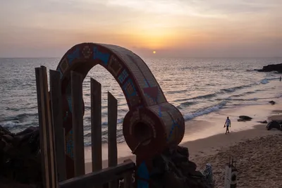 Pastel sunset at Toubab Diallaw, Senegal: the sun nearly touching the water, a weathered gate in the foreground, and a man walking alone along the shore